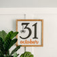 A square-shaped Halloween sign with the date '31 October' written in black, with a spider hanging from the number 1. The sign has a white background and a weathered walnut stain.